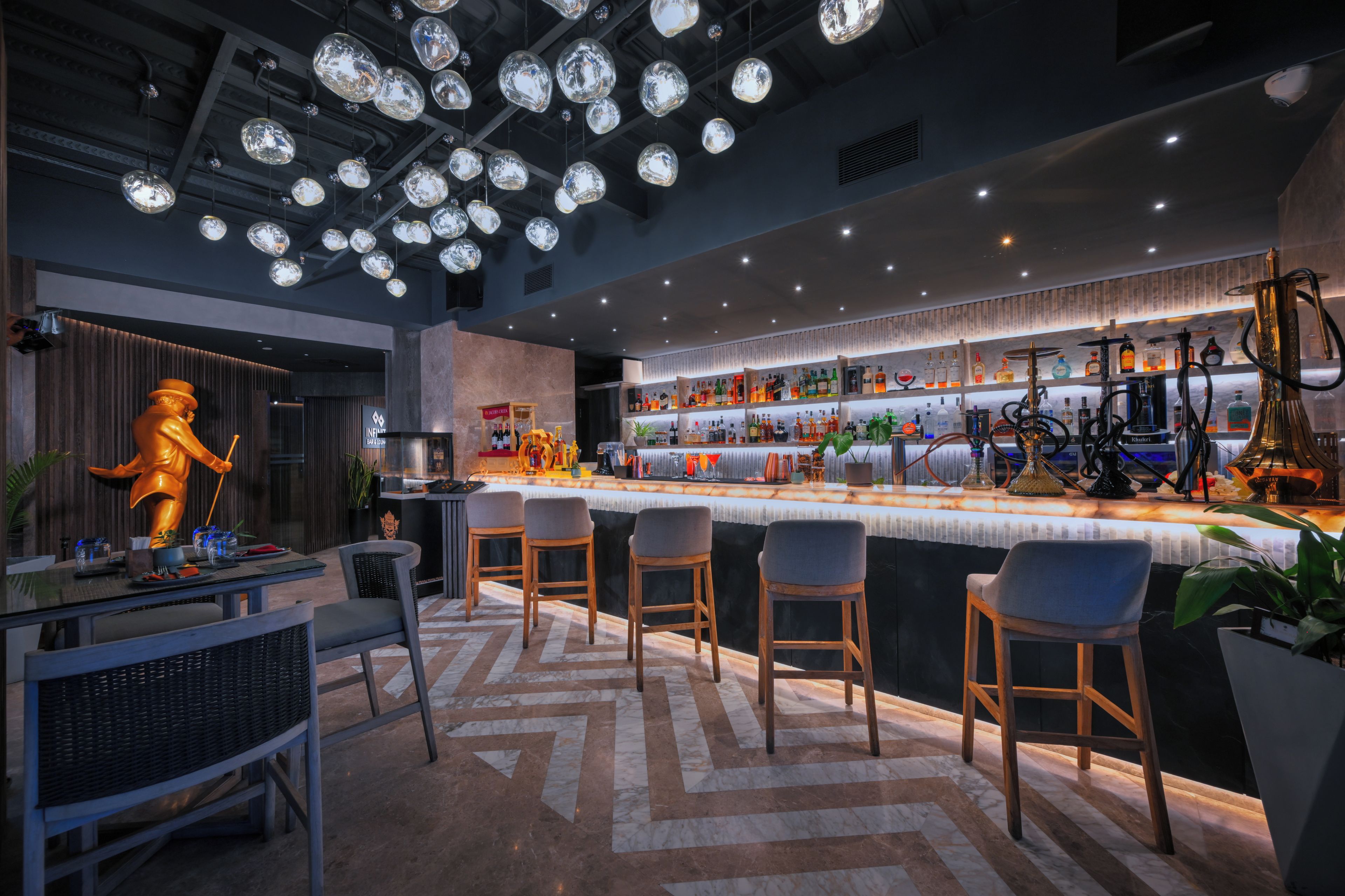 A modern bar area with a high ceiling and pendant lighting, featuring a bar counter with stools and a variety of bottles.