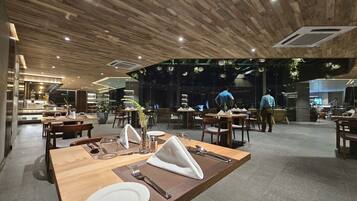 A modern restaurant interior with wooden ceiling panels, tables set with white plates, and a bar area visible in the background.