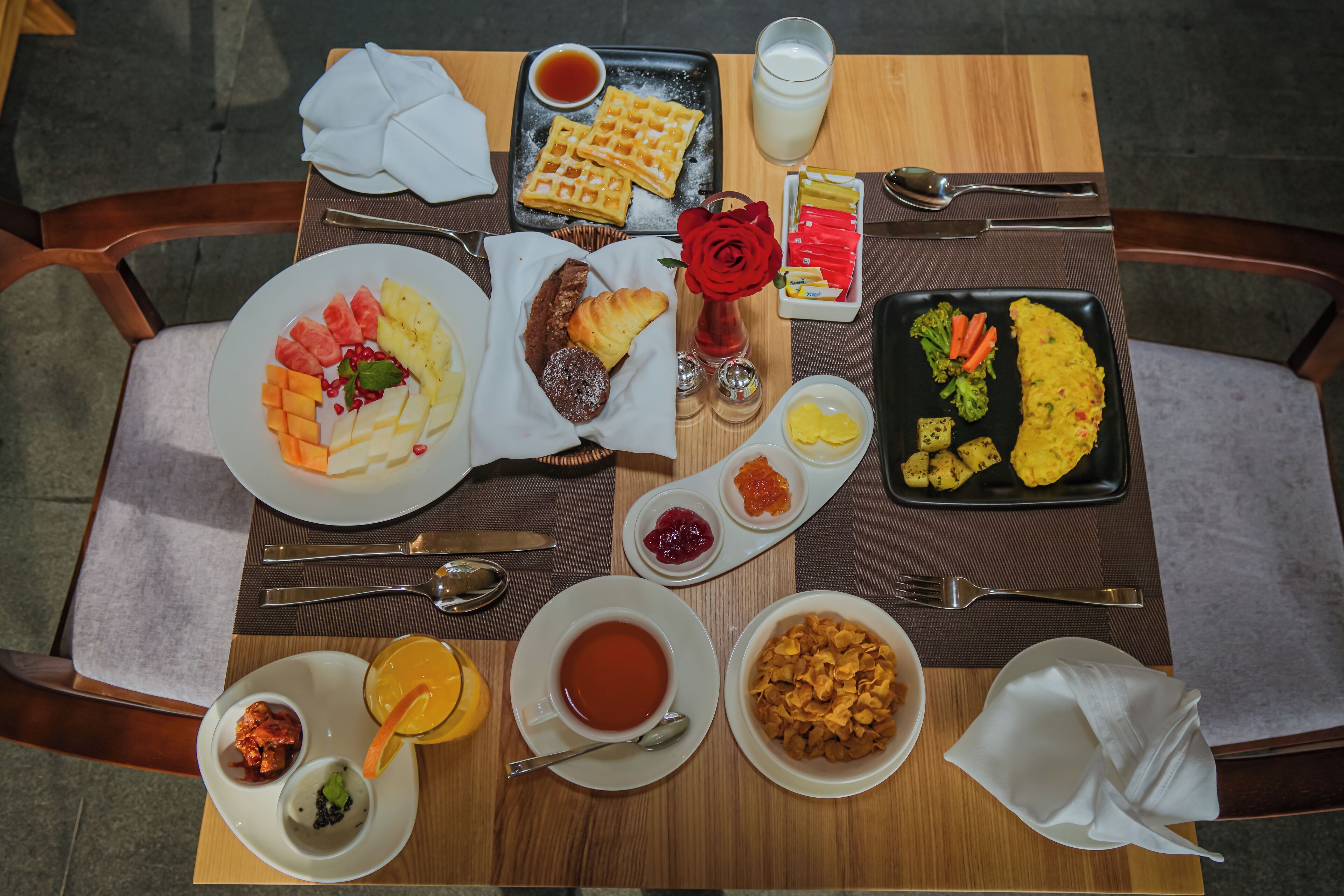 Una mesa de desayuno con diferentes platos, como waffles, fruta y una rosa roja.