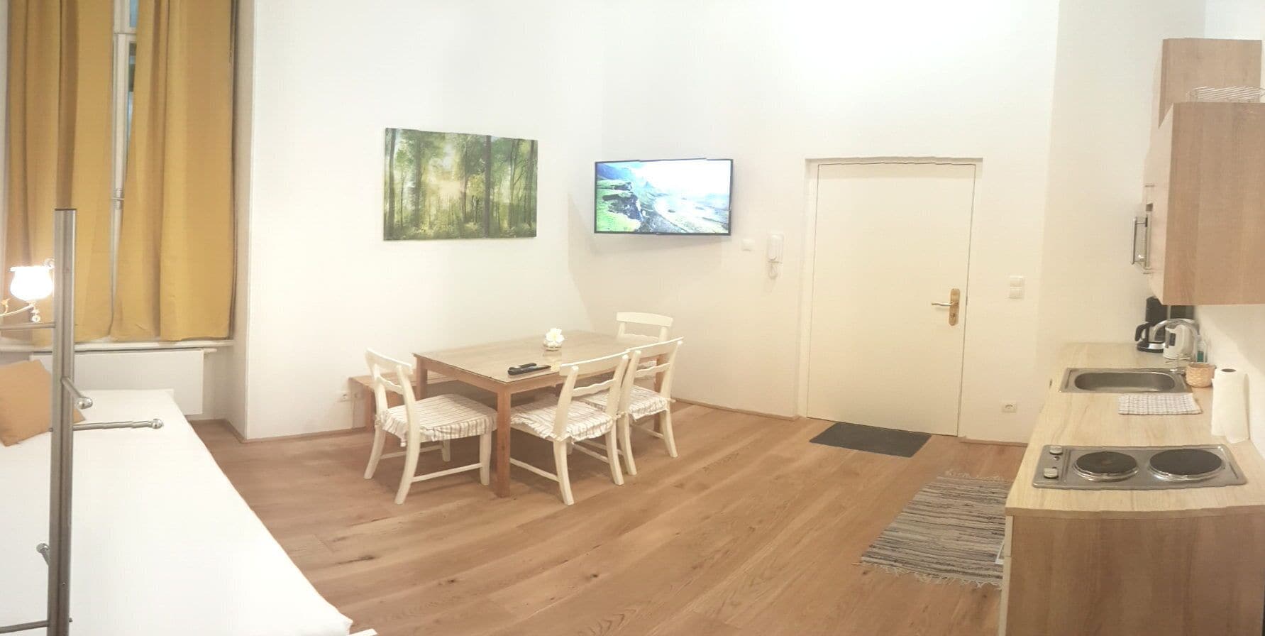 A compact room with a dining table, chairs, a TV, a door, and a kitchenette.