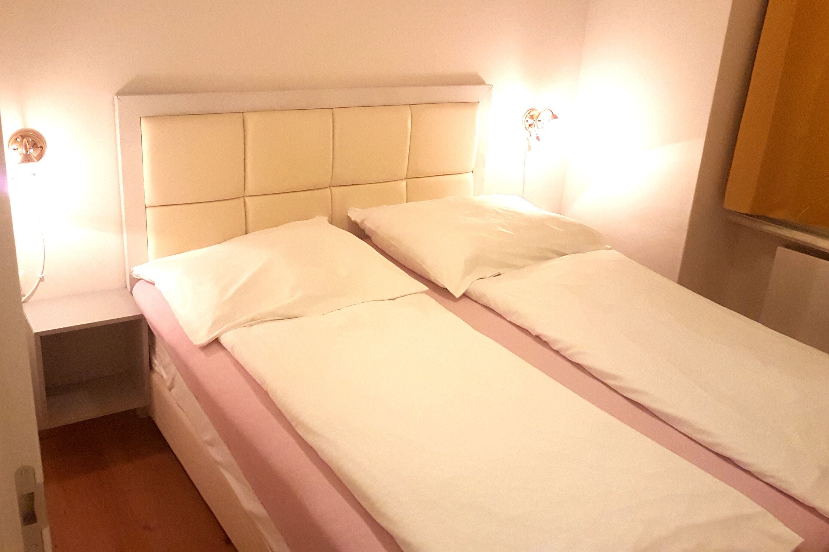 Ein Doppelbett mit gepolstertem Kopfteil, ein Nachttisch und eine an der Wand befestigte Lampe.