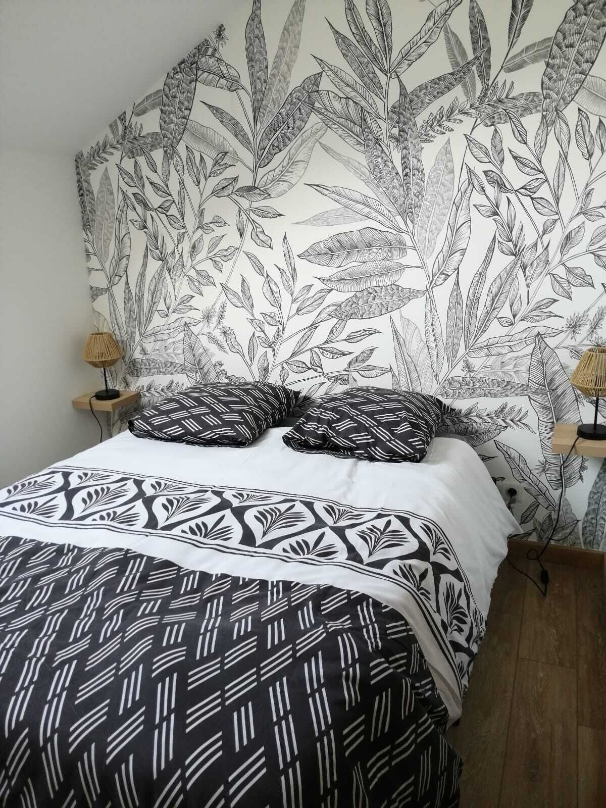 Un lit avec une couvre-lit à motifs et des oreillers, un mur recouvert d’un papier peint à motifs végétaux, et deux lampes de chevet.