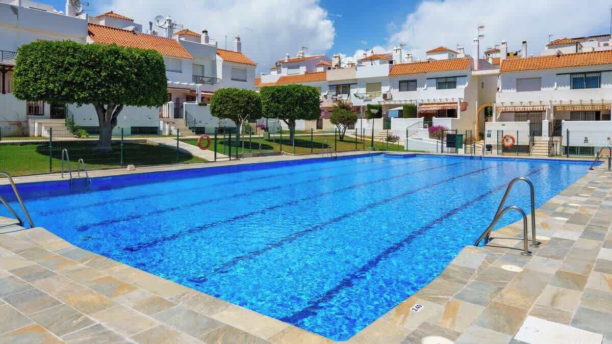 A swimming pool with tiled edges, surrounded by white buildings with orange roofs, and small trees in the foreground.