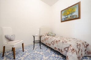 A small bedroom with a single bed, a chair, a small table, and a painting on the wall. - Villa Carrino 2 (Positano)