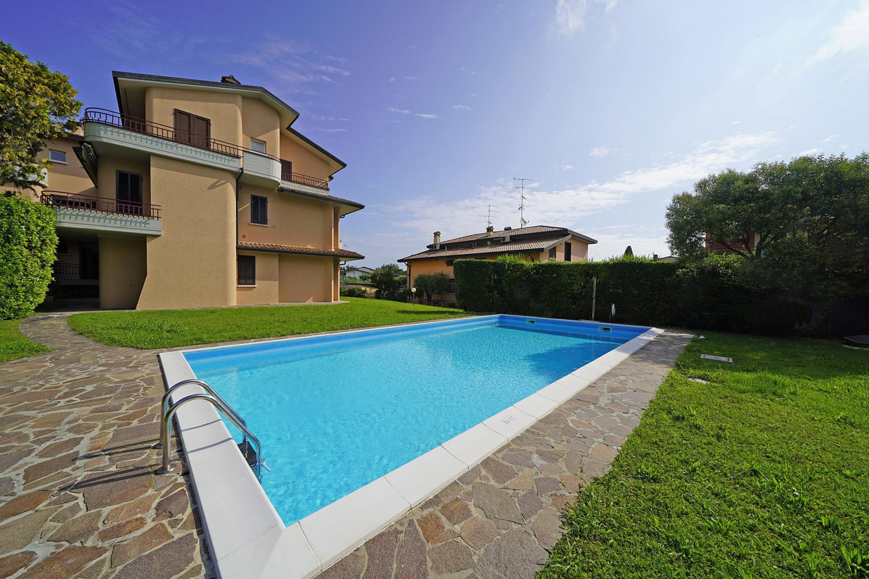 Une piscine aux bords recouverts de pierres, entourée d’une pelouse bien entretenue et d’un immeuble à plusieurs étages doté de balcons.