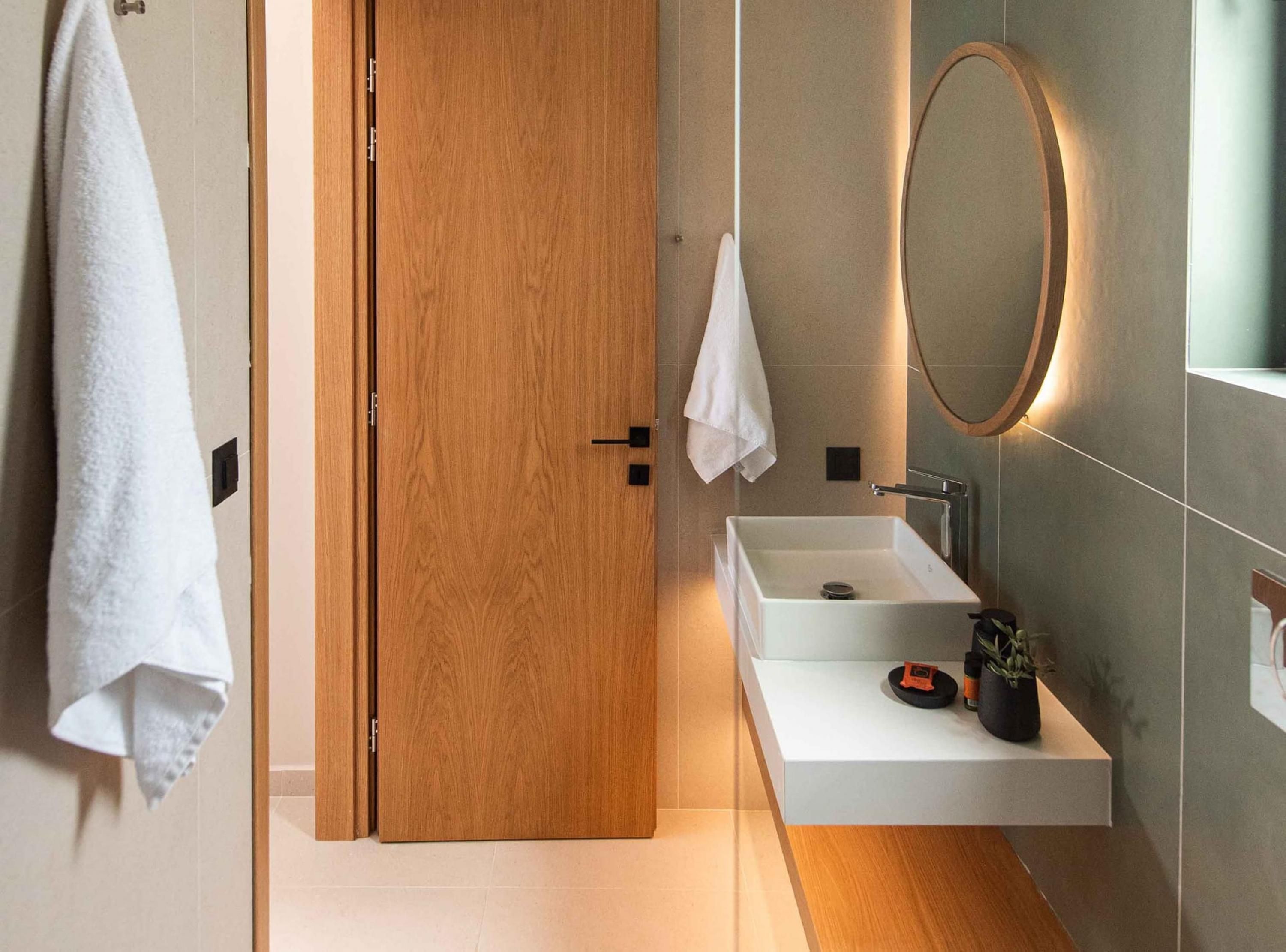 Une salle de bain moderne avec une porte en bois, un lavabo blanc, un miroir et une serviette accrochée au mur.