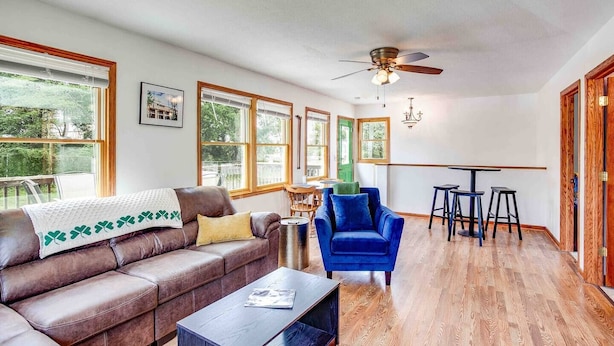 A spacious living area with a brown leather sofa, a blue armchair, and a wooden coffee table. There are large windows providing natural light and a view of the outdoors.