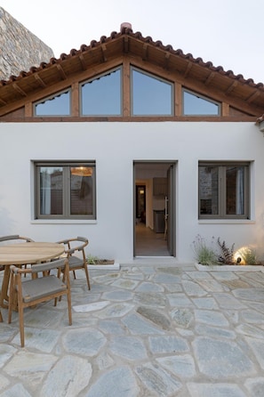 A modern house with a stone patio, wooden furniture, and a kitchen visible through an open door.