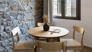 A dining area with a round wooden table, four wicker chairs, a woven pendant light, a stone wall, and a window with a view of a stone building. - Lichnos Luxury Suites Stoa (Almyros)