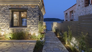 A stone building with a tiled roof, a window with a dark frame, and a pathway leading to a gate. - Lichnos Luxury Suite (Almyros)