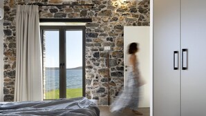 A bedroom with a stone wall, a bed with a grey blanket, a sliding glass door to a balcony, and a white wardrobe. - Lichnos Luxury Suite (Almyros)