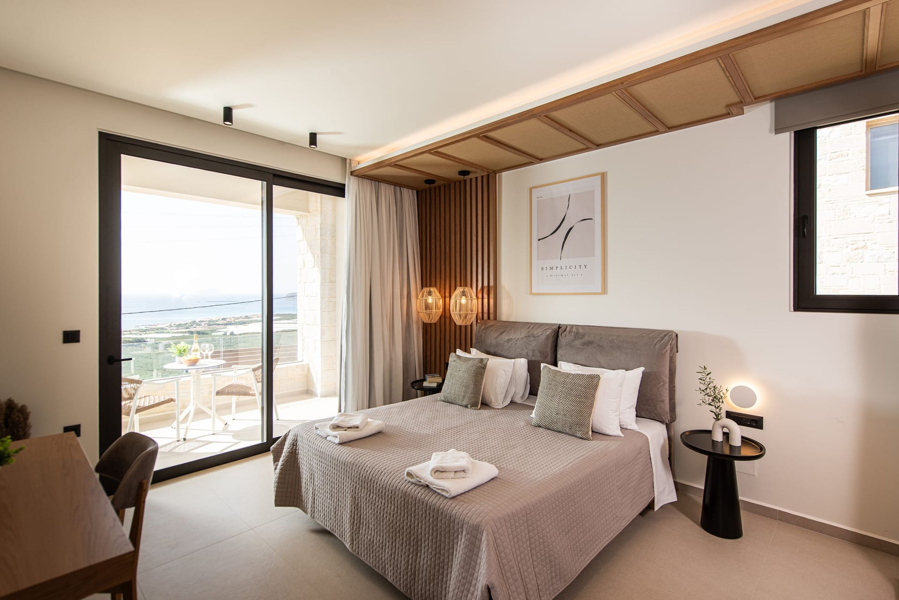 A modern bedroom with a large bed, a bedside table, a small round table with a lamp, a framed picture on the wall, and a sliding glass door leading to an outdoor area.