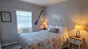 Ein Schlafzimmer mit einem Bett, zwei Nachttischen mit Lampen, einem Fenster mit Jalousien, einer Topfpflanze und einer Deckenleuchte.