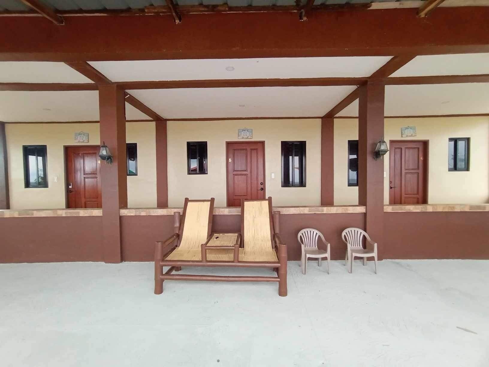 A covered outdoor seating area with a wooden bench and two plastic chairs, a wall with several windows, and a door.
