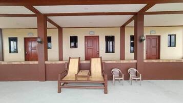 A covered outdoor seating area with a wooden bench and two plastic chairs, a wall with several windows, and a door.