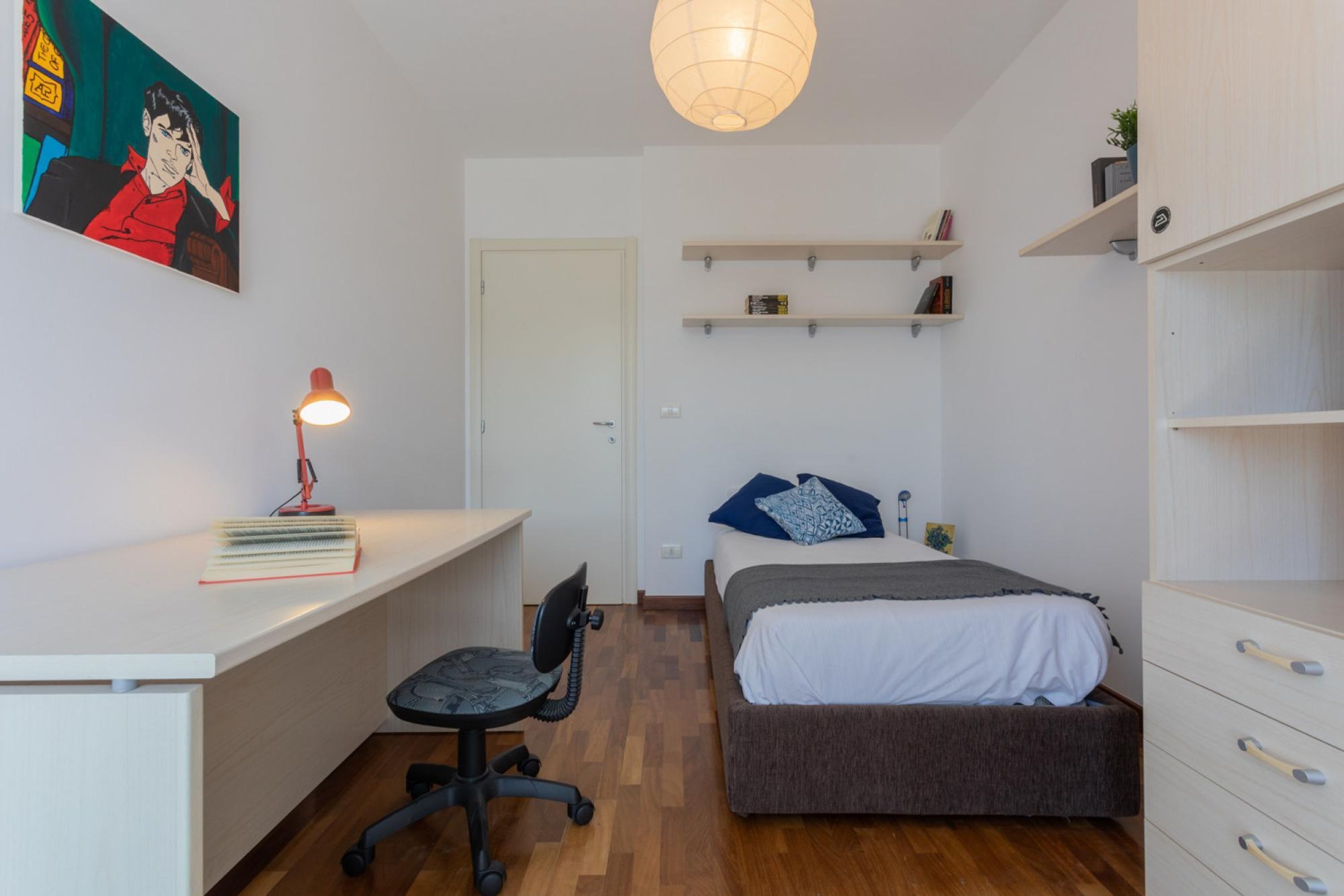A bedroom with a bed, a desk, a chair, a hanging light, and shelves with books and decorative items.
