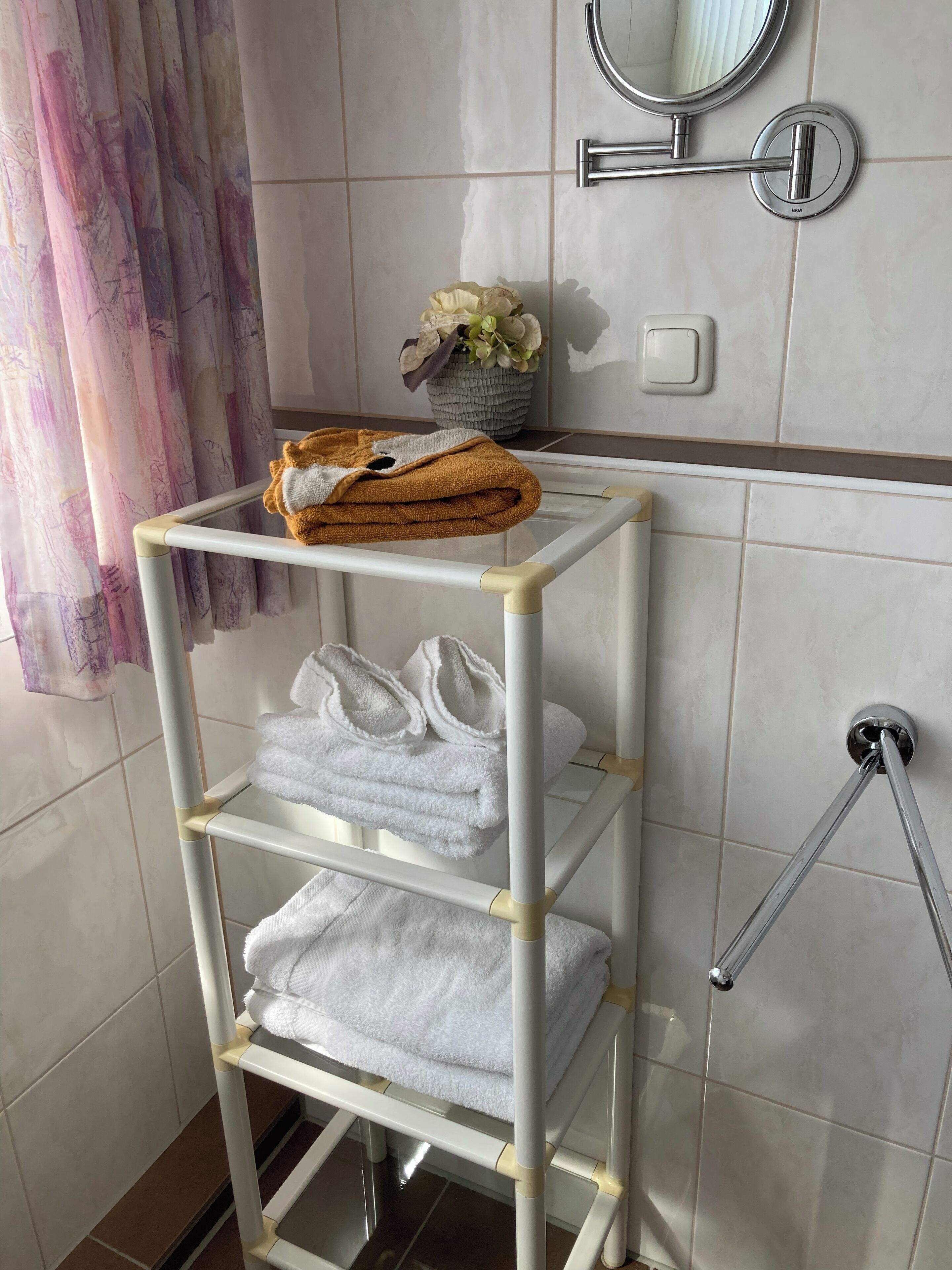 Une salle de bain avec un porte-serviettes blanc sur lequel sont pliées des serviettes, et une serviette marron posée sur une étagère blanche.