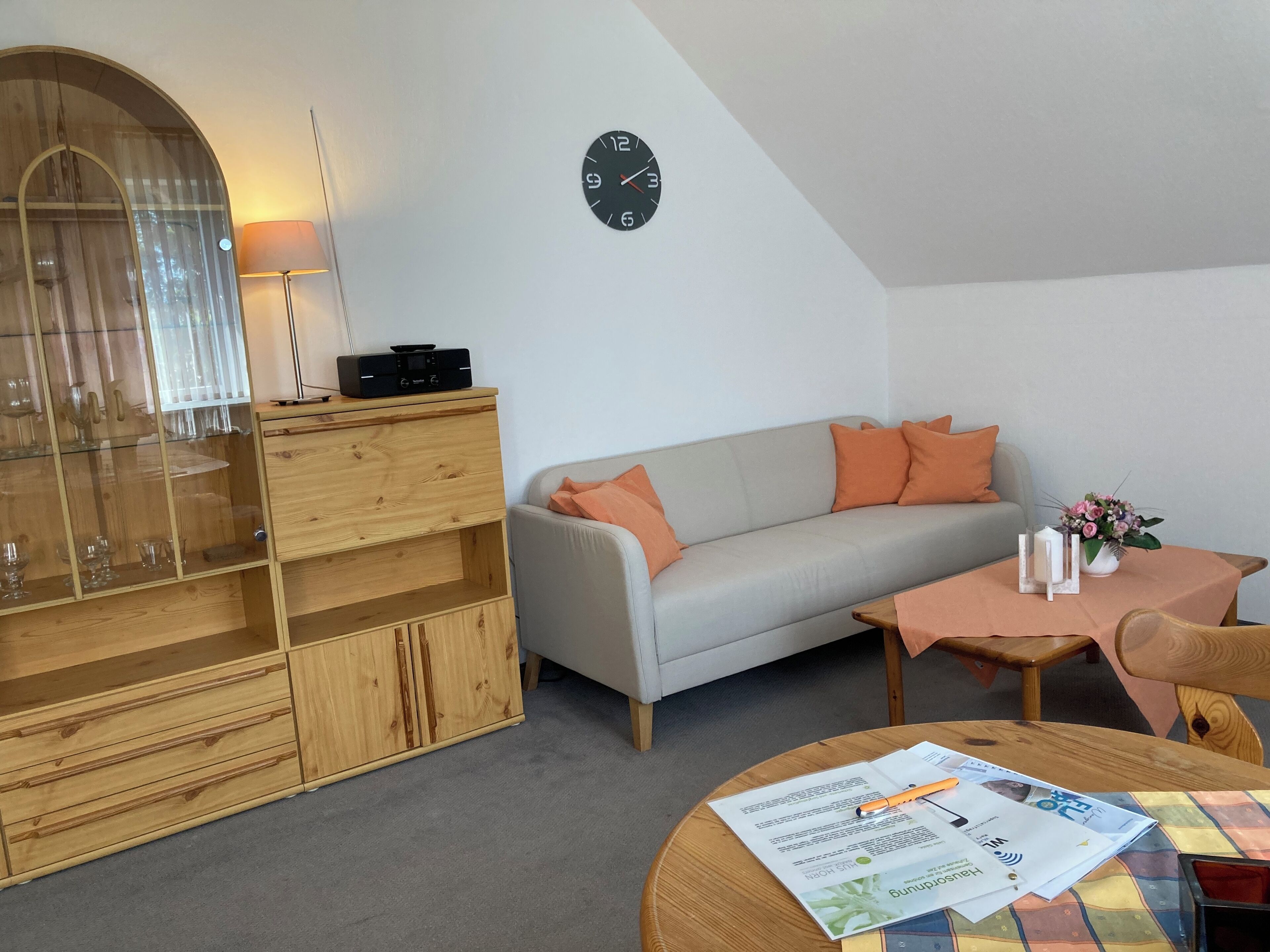 Un salon moderne avec un canapé, un meuble en bois, une table ronde sur laquelle se trouvent des papiers et un stylo, et une horloge au mur.