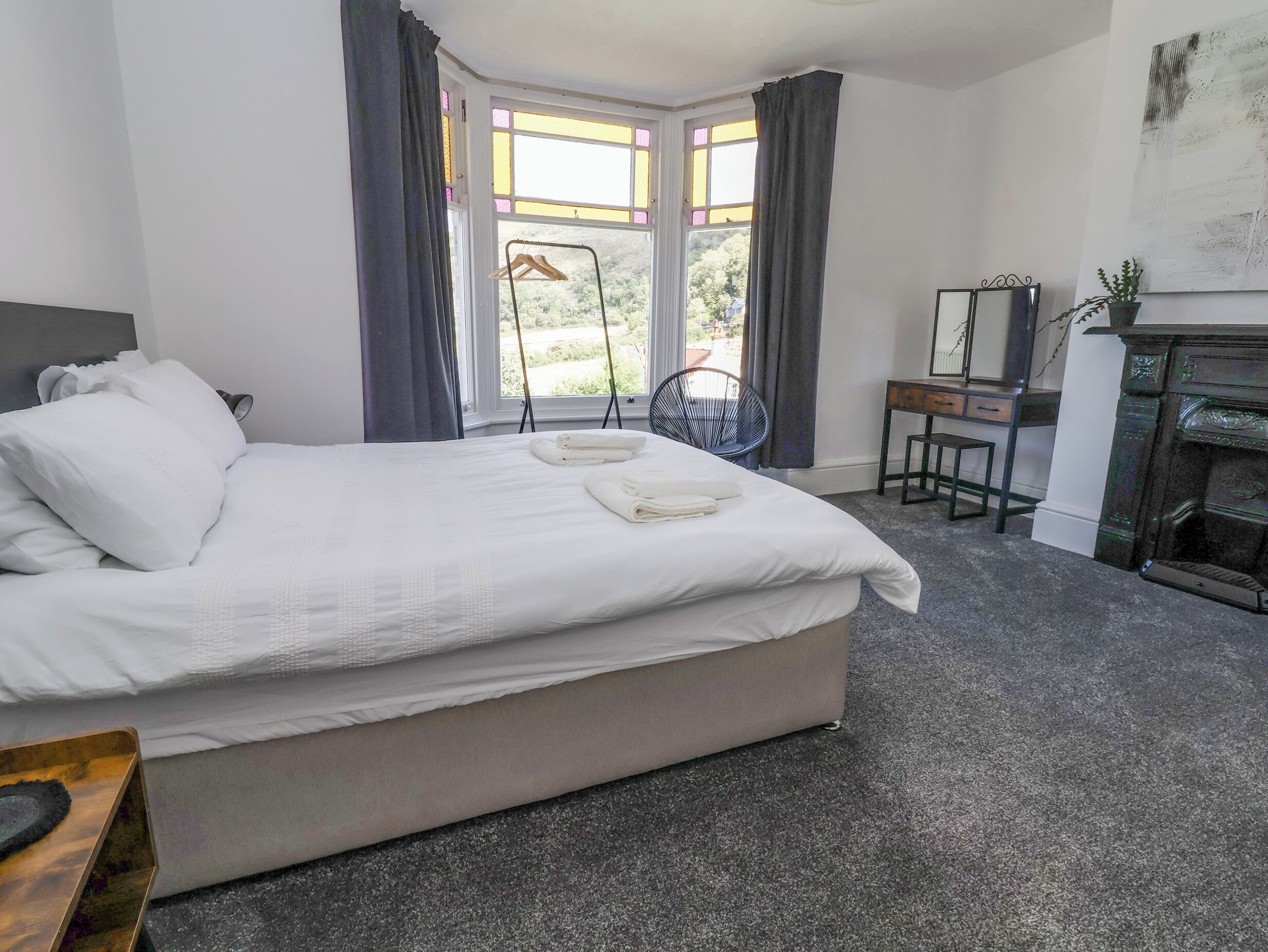 A neatly made bed with white linens, a small wooden side table, a chair, and a fireplace.