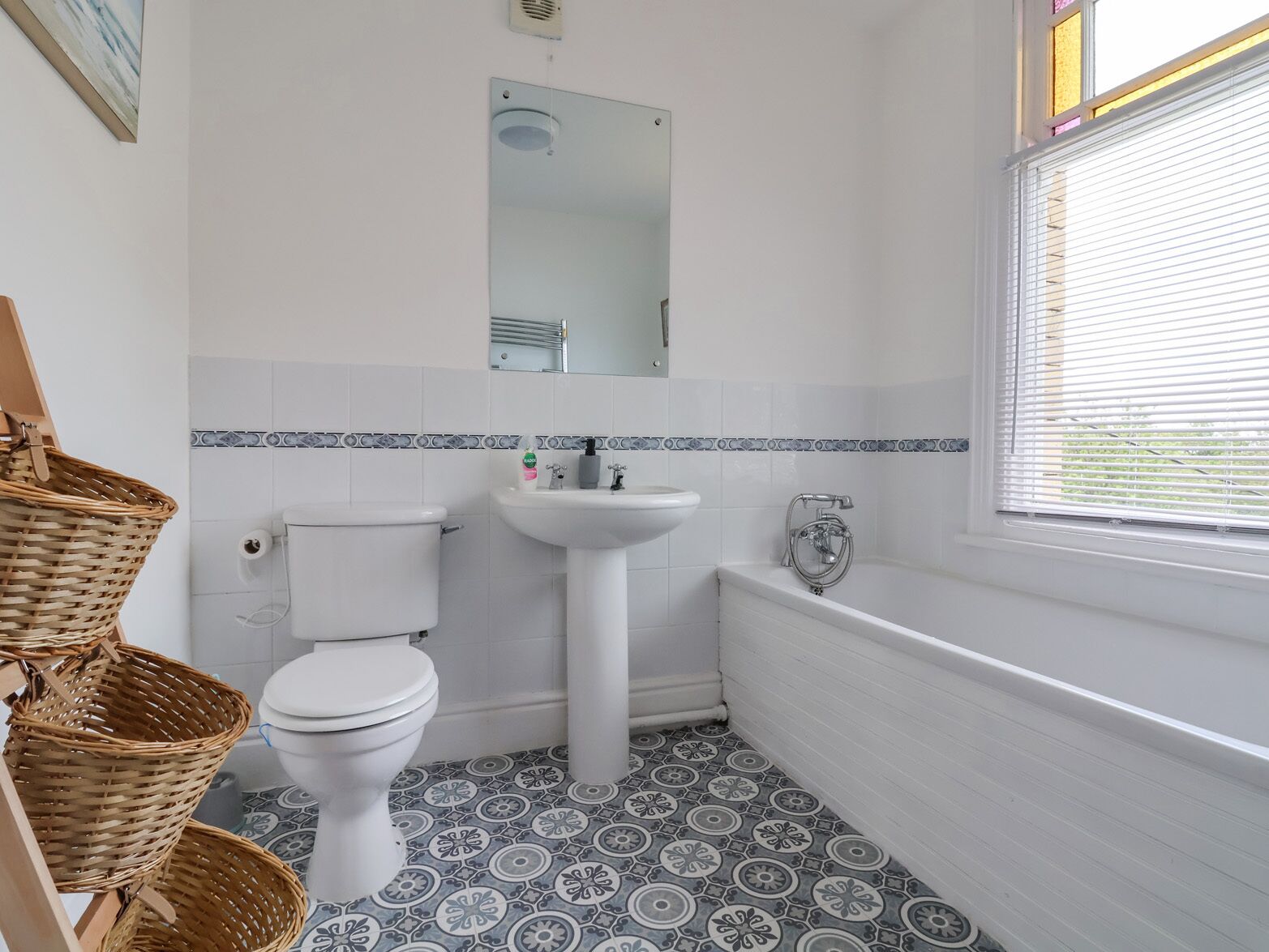 A bathroom with a toilet, sink, and bathtub. There are wicker baskets and a window with blinds.