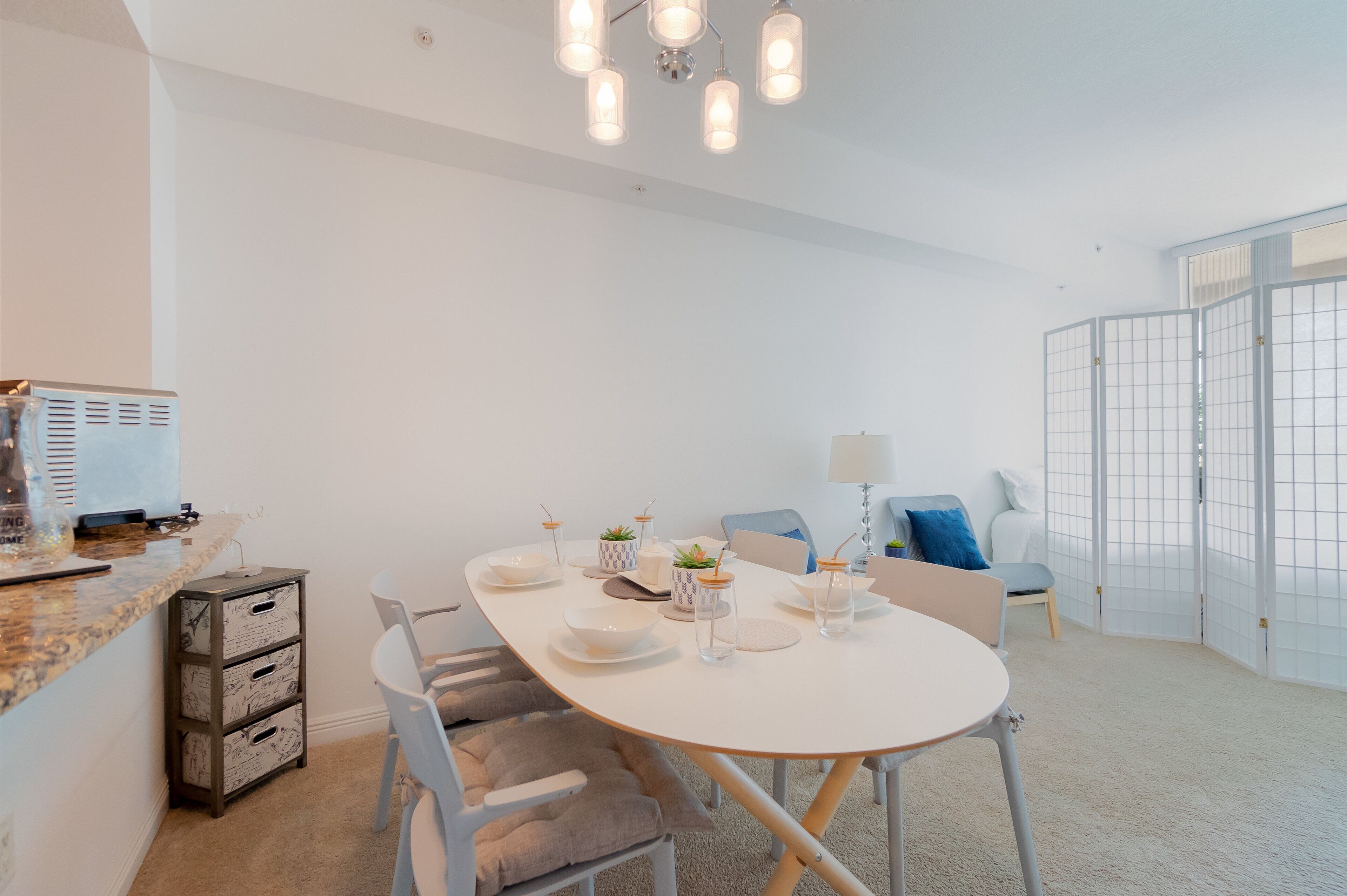 A modern dining area with a white table set for four, surrounded by white chairs. There is a kitchen area with a countertop and cabinets to the left. A partition with a grid pattern separates the dining area from a seating area with a blue chair and a lamp.