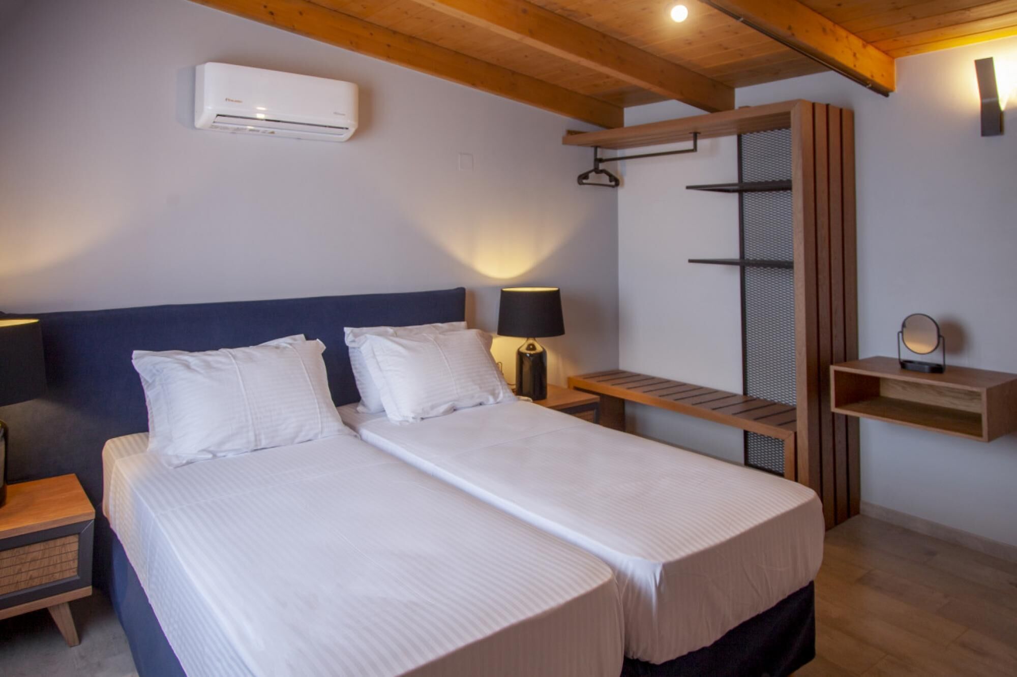 A double bed with white linens, a bedside table with a lamp, a wall-mounted air conditioner, and a wooden shelf with a mirror.