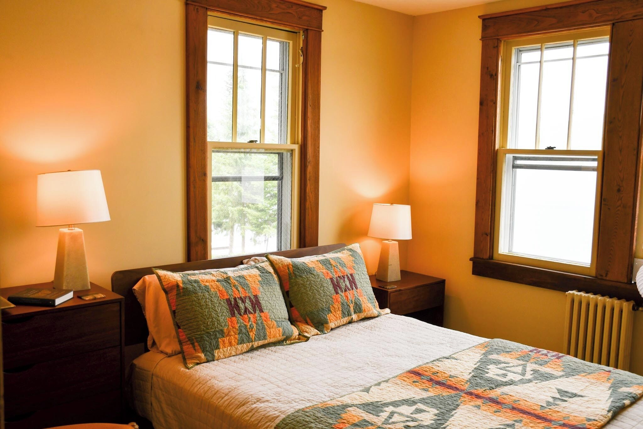 A bedroom with a bed, two nightstands, two lamps, and two windows.