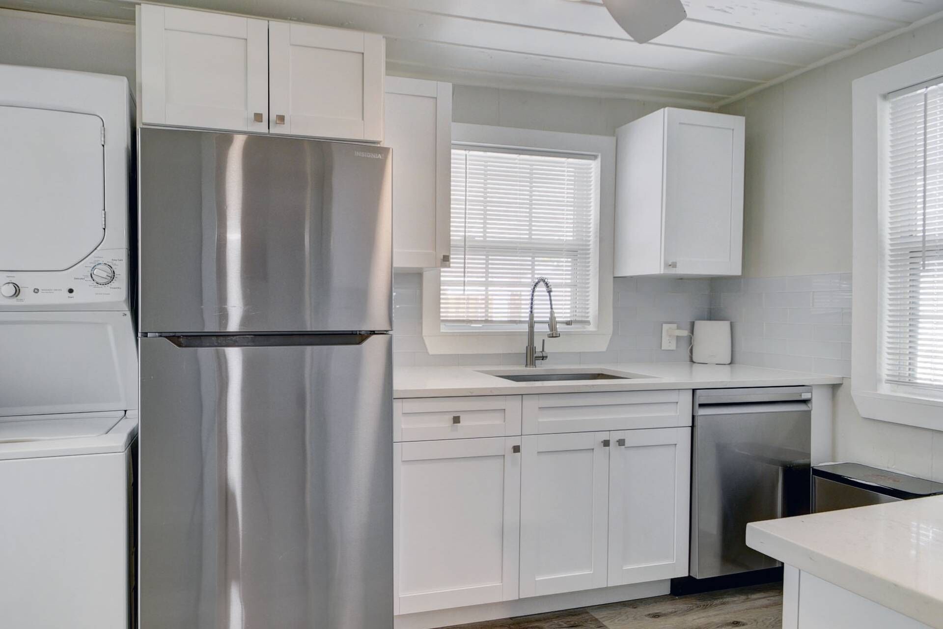 A modern kitchen with white cabinetry, a stainless steel refrigerator, a dishwasher, and a window with blinds.
