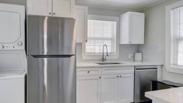 A modern kitchen with white cabinetry, a stainless steel refrigerator, a dishwasher, and a window with blinds.