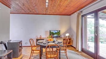 A dining area with a round table, chairs, a television, and a view of greenery through a sliding door.