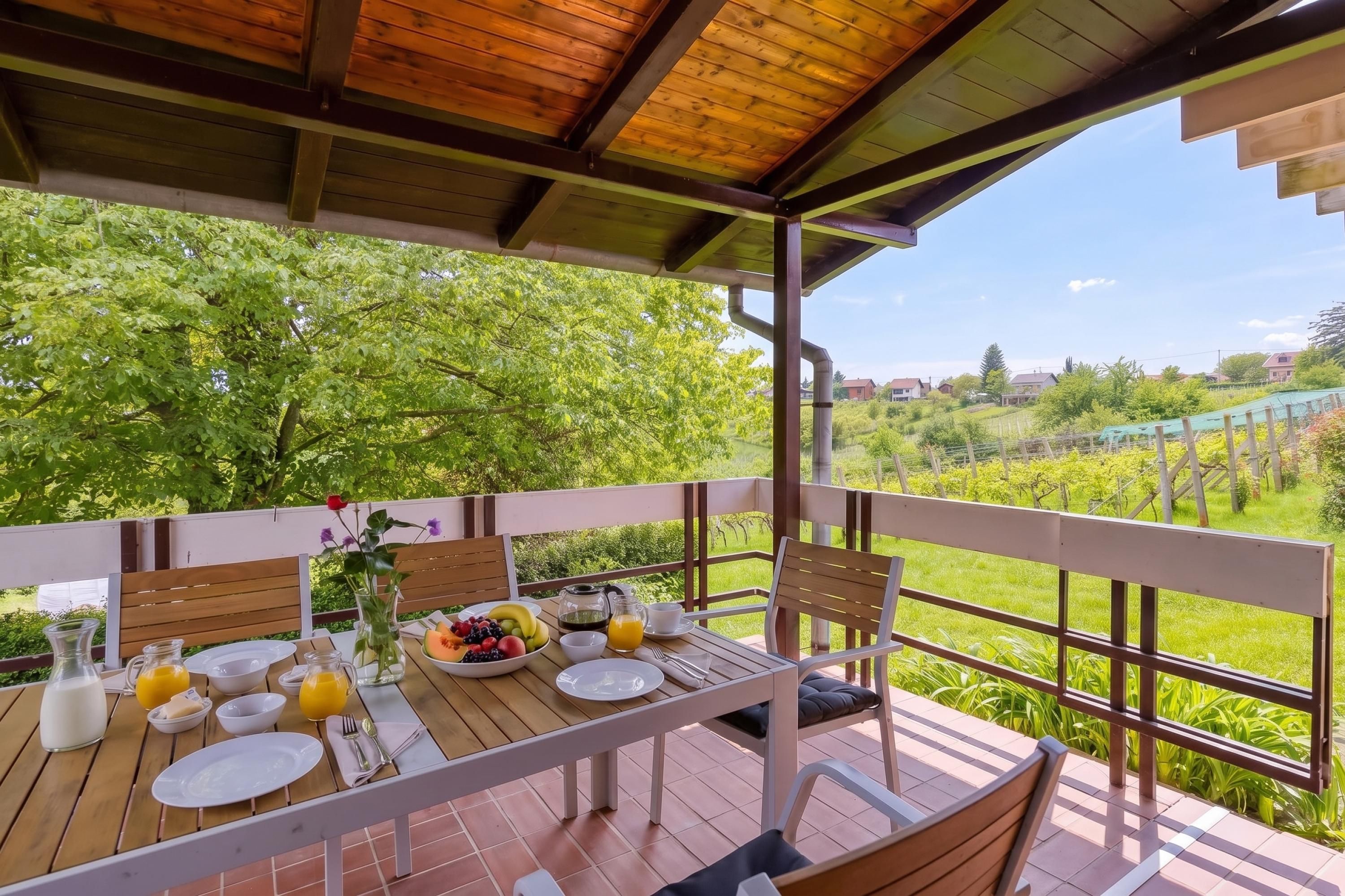 Deck de madeira com mesa posta para café da manhã, com vista para um jardim exuberante e edifícios ao longe.