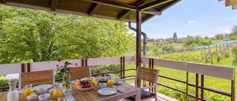 Deck de madeira com mesa posta para café da manhã, com vista para um jardim exuberante e edifícios ao longe.