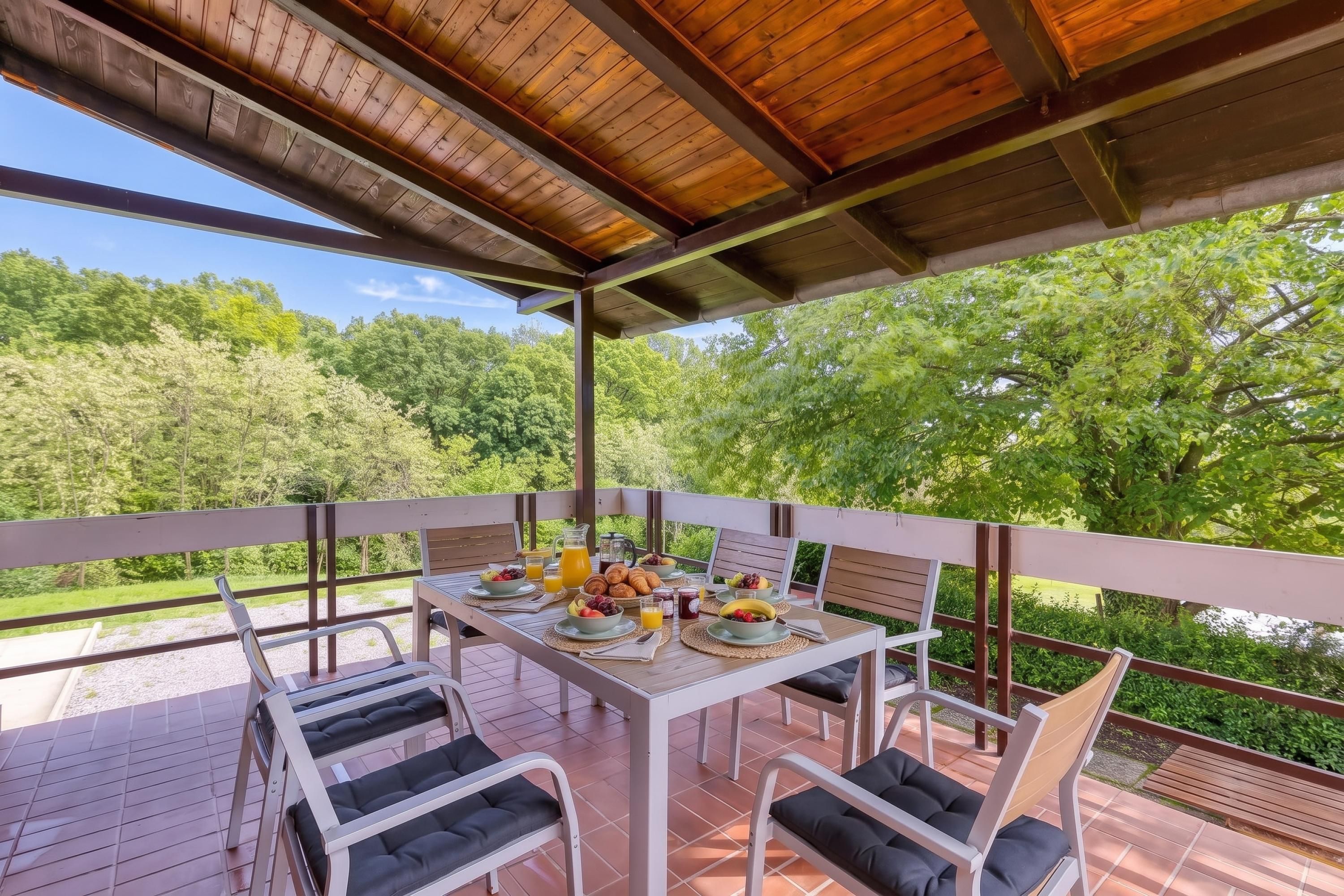 Un espacio de comedor al aire libre techado, con una mesa dispuesta para el desayuno, rodeada de sillas y con vistas a un paisaje boscoso.