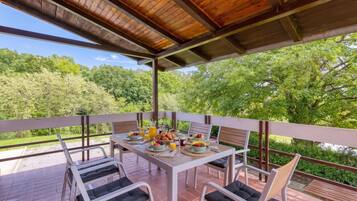 Un espacio de comedor al aire libre techado, con una mesa dispuesta para el desayuno, rodeada de sillas y con vistas a un paisaje boscoso.