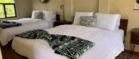 Habitación de hotel con dos camas, cada una con ropa blanca y almohadas decorativas. Un plaid estampado cubre la cama en primer plano. El piso es de baldosas de piedra y hay un ventanal con vistas a la vegetación.