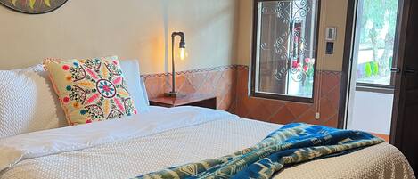 A neatly made bed with a patterned blanket, a decorative pillow, a wall clock, and a window with a view of greenery.