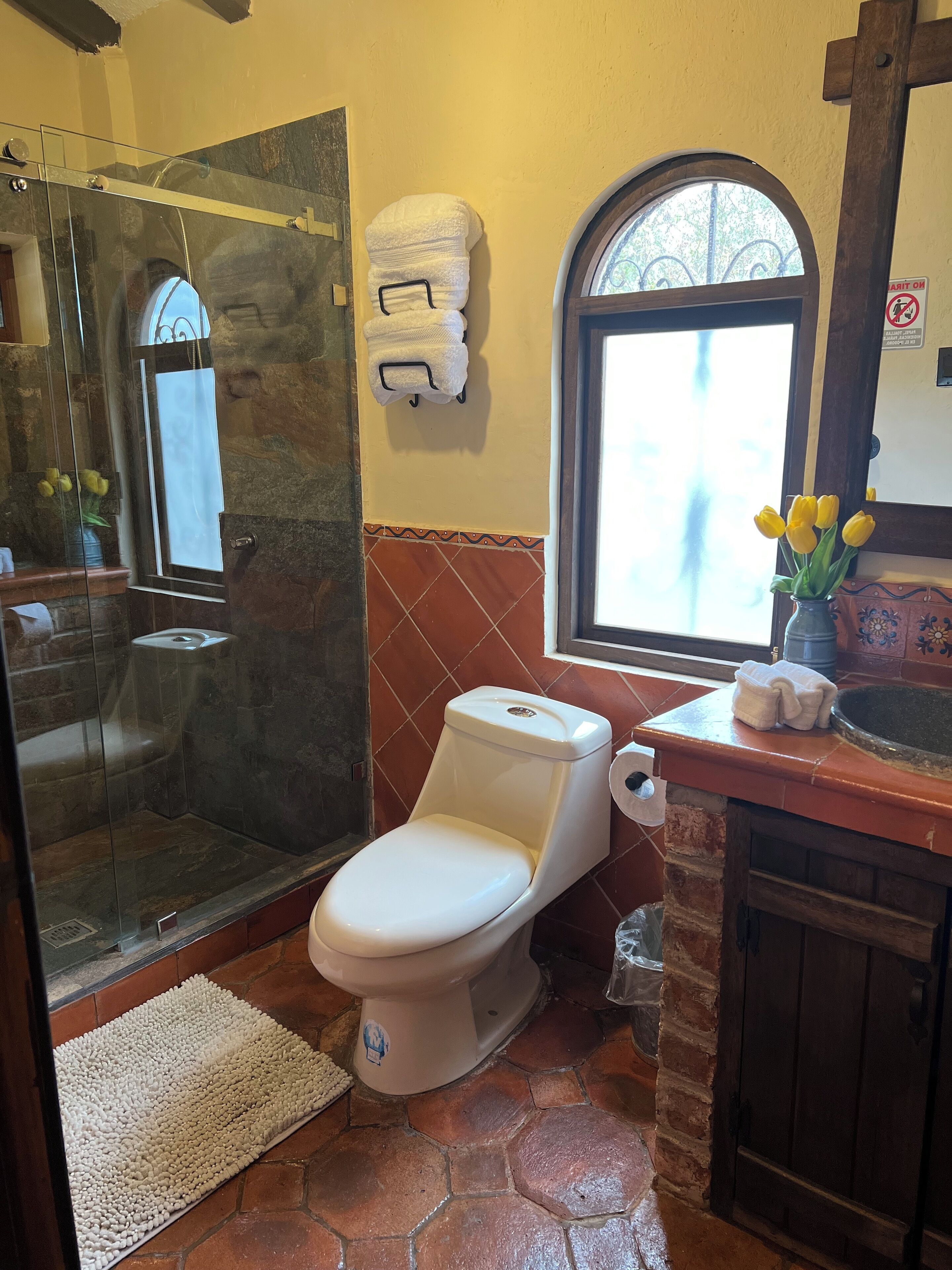 Un baño con ducha de vidrio, un inodoro, un lavamanos con un florero de flores amarillas y una ventana con vistas a árboles.