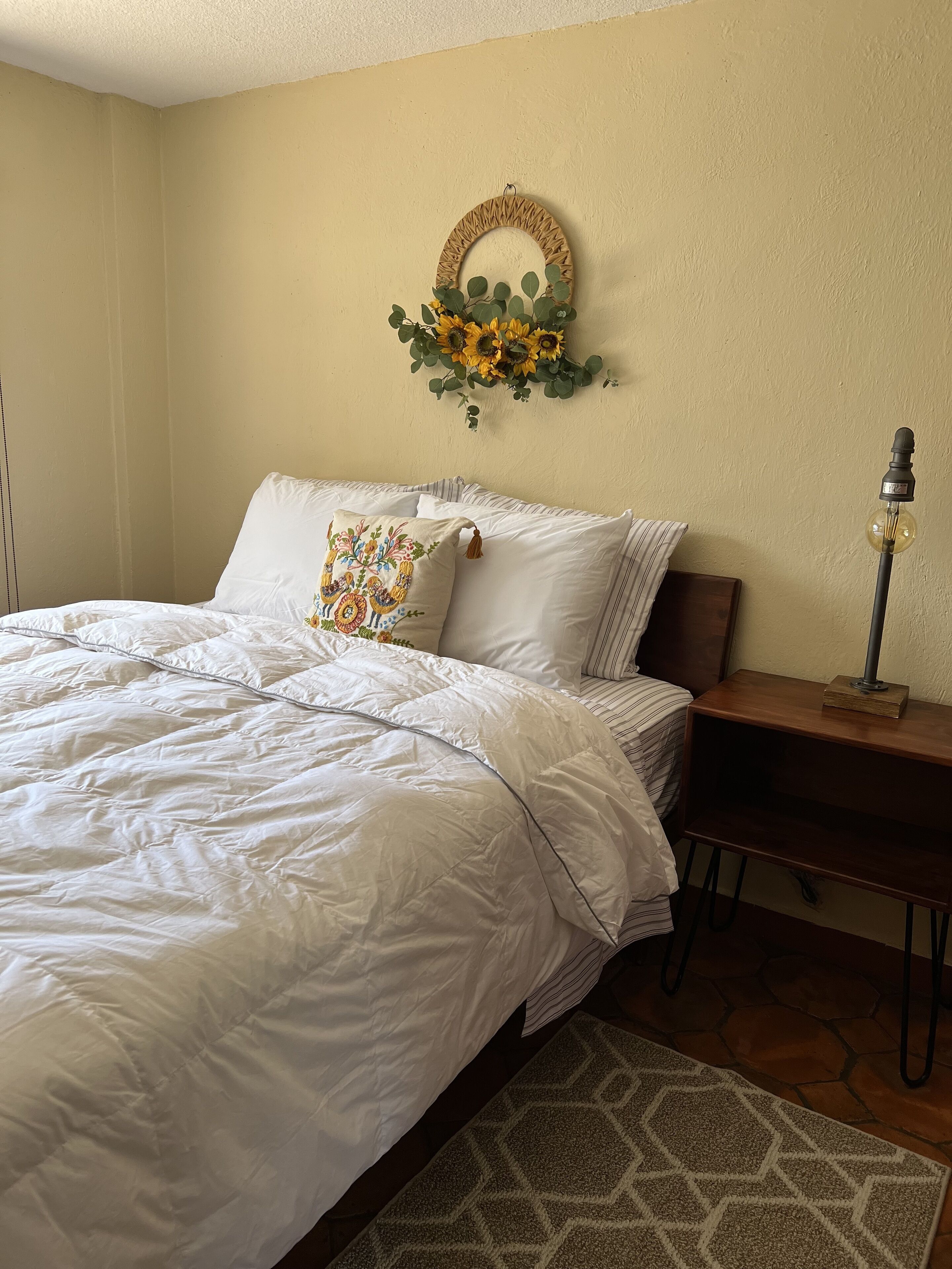 Un lit bien fait, avec une literie blanche et un coussin décoratif, une table de chevet avec une lampe et un miroir mural orné d’un motif de tournesol.
