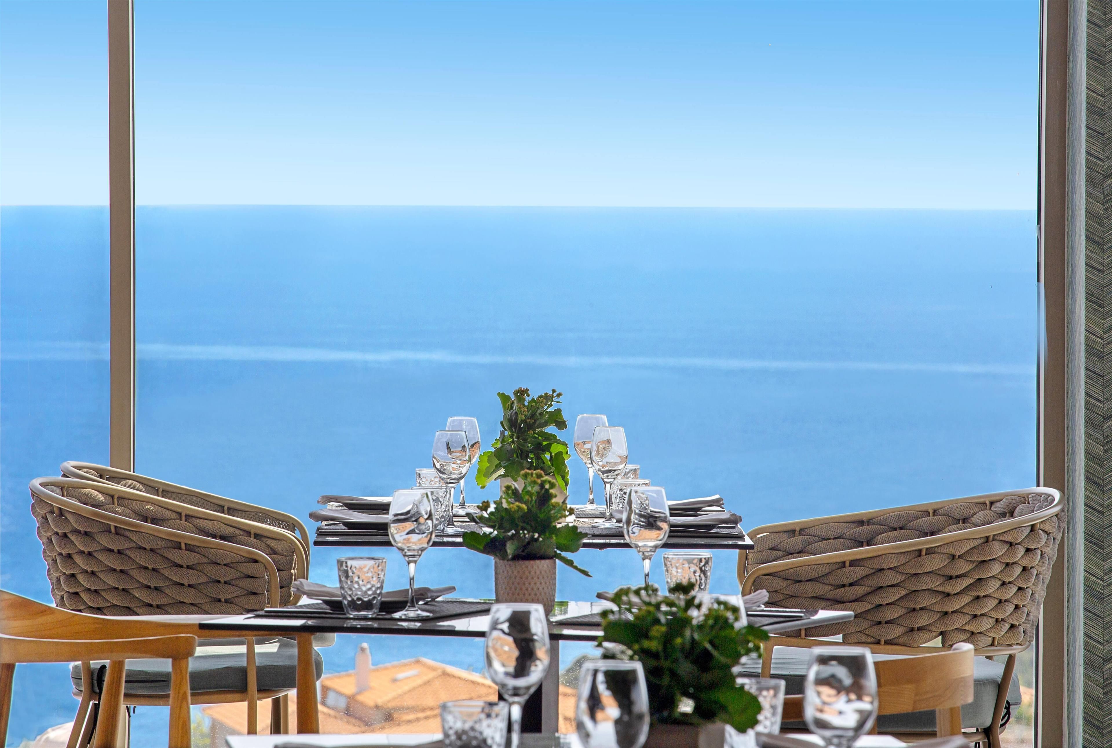A dining table set with chairs, a potted plant, and a view of the sea.