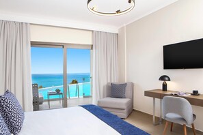 A hotel room with a bed, a desk, a chair, a TV, and a balcony with a view of the ocean. - Ajul Luxury Hotel & Spa Resort Registry Collection (Kassandra)