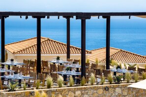An outdoor dining area with tables and chairs, a stone wall, and a view of the sea. - Ajul Luxury Hotel & Spa Resort Registry Collection (Kassandra)
