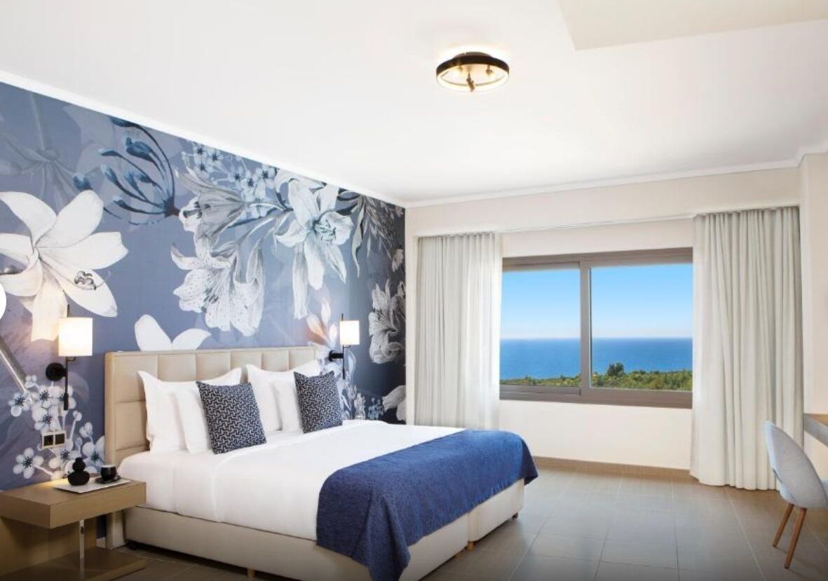 A bedroom with a large bed, a nightstand, a lamp, a view of the ocean, and a window with curtains.