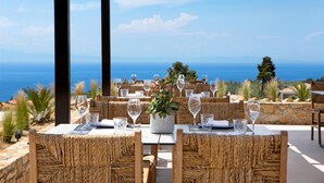An outdoor dining area with wicker furniture, a table with a vase of flowers, and a view of the sea. - Ajul Luxury Hotel & Spa Resort Registry Collection (Kassandra)