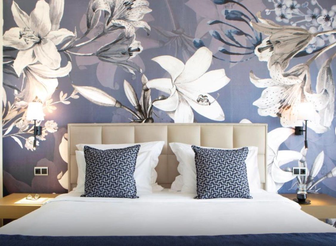 A hotel room with a large bed, two patterned pillows, a headboard, a nightstand, a lamp, and a floral wall mural.
