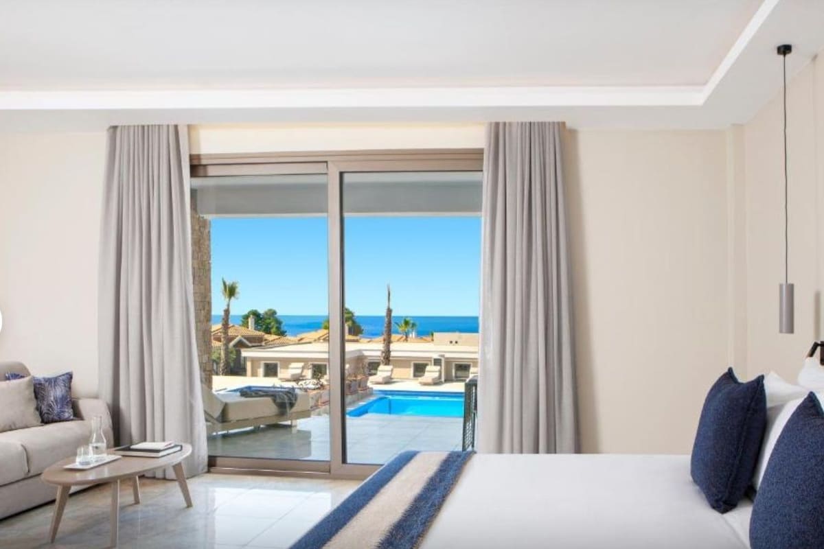 A hotel room with a bed, a sofa, a coffee table, and a view of a pool and ocean.