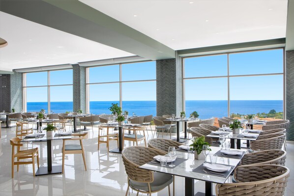 A restaurant with tables and chairs arranged for dining, large windows offering a view of the ocean, and a modern interior design. - Ajul Luxury Hotel & Spa Resort Registry Collection (Kassandra)
