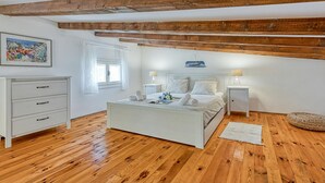 Une chambre avec un plancher en bois, un lit recouvert de draps blancs, une table de chevet avec une lampe, une commode et un tableau accroché au mur.
