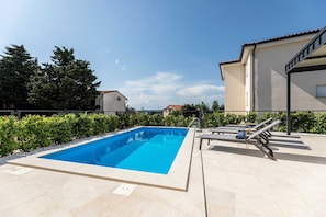 Una piscina rectangular con solárium empedrado, rodeada de setos bajos y un cielo despejado.
