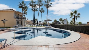 Une piscine avec des chaises longues, des palmiers et un bâtiment doté d’un toit en tuiles.