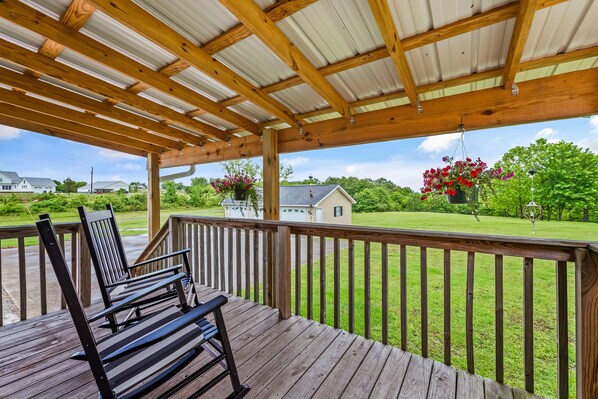 Une terrasse en bois surmontée d’une pergola, donnant sur une cour verdoyante avec une maison et un pot de fleurs suspendu.